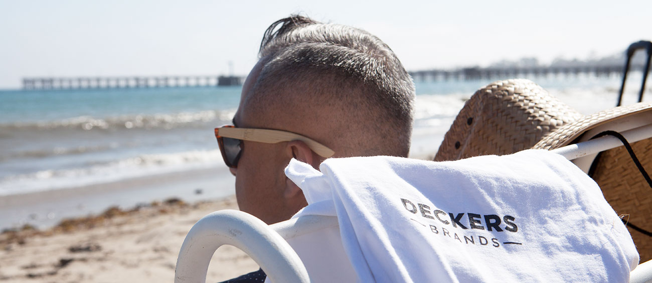 Man sitting at the beach with Deckers Brands shirt draped over chair.