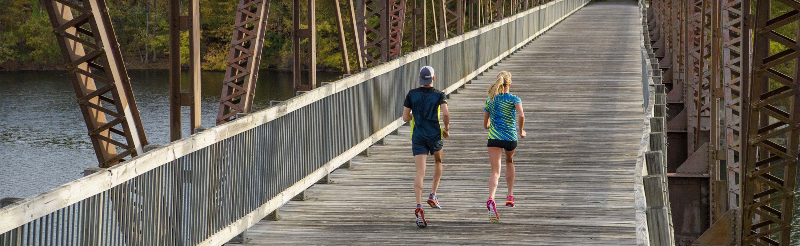 Man and woman running across a bridge wearing Hoka One One running shoes.